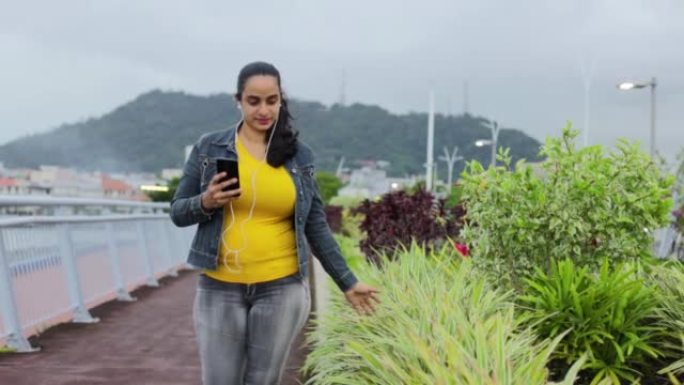 一个拉美裔女孩带着电话和有线耳塞在巴拿马植物附近行走的高清视频
