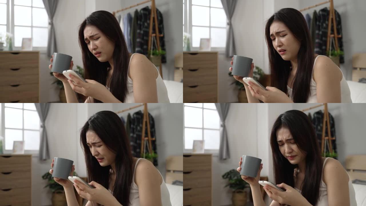 拿着早茶的亚洲女孩在家里的卧室里用做鬼脸在手机上浏览可怕的信息时摇头并向下滚动屏幕