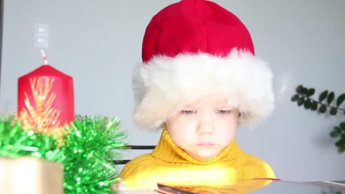 一个穿着黄色毛衣和红色圣诞老人帽子的小孩看着平板电脑，突然抬头看着坐在桌子旁的相机。