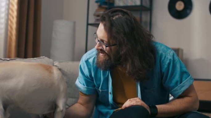 一个男人和他的四条腿的毛茸茸的朋友进入客厅。一个戴着眼镜的大胡子男人正在和狗开玩笑地抚摸它。宠物咬住