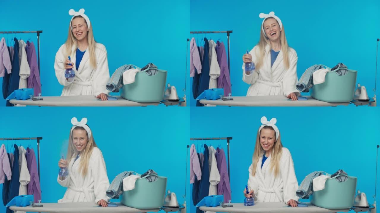一个穿着浴袍的微笑金发女郎早上正在洗衣服，熨烫，拿着一瓶喷水。工作室蓝色背景上的家庭主妇肖像