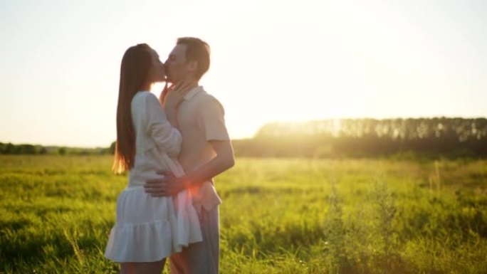 幸福的年轻夫妇在夏日田野的日落背景下拥抱、亲吻和牵手。草地户外浪漫约会爱情。