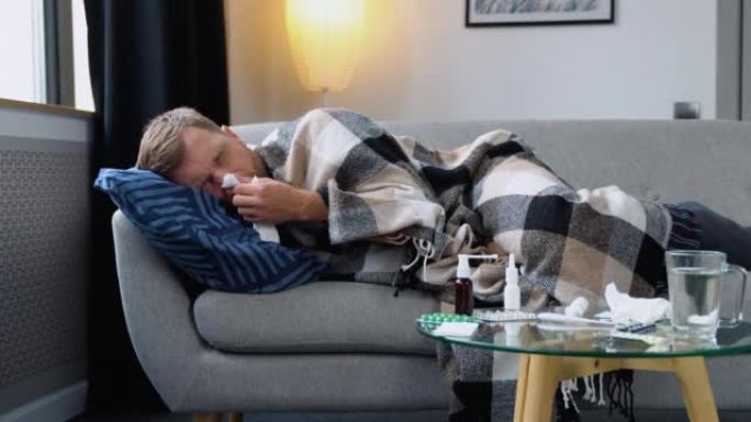 季节性疾病时期。特写一名患有流感、发热和头痛的病人躺在沙发上的格子里。一个男人流鼻涕和普通感冒。病毒