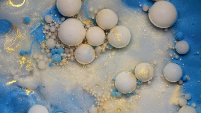 彩色白色和金色油漆气泡，金色灰尘散布在蓝色水彩表面上，破裂并混合在抽象设计中。闪闪发光的颗粒，墨球滴