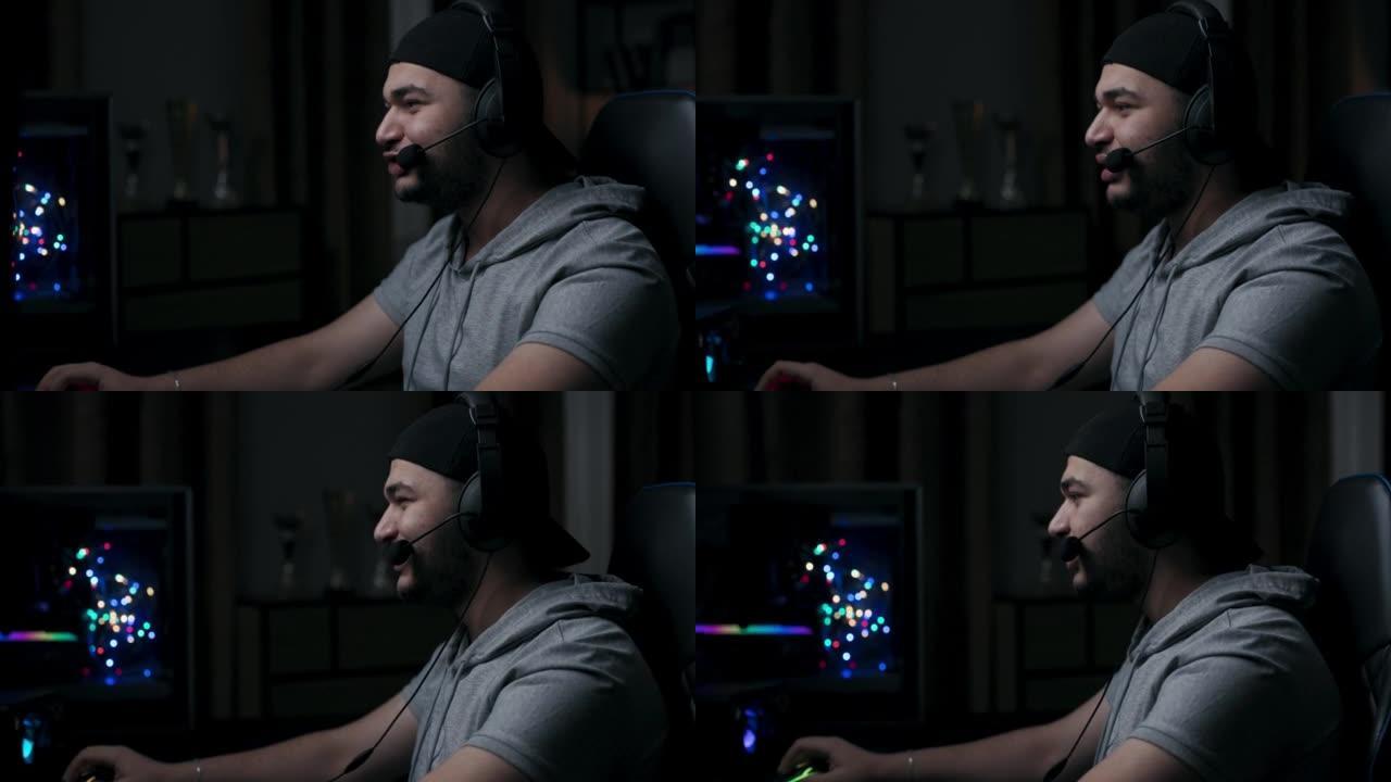 穿着灰色t恤和黑帽子的胡子的家伙正在玩网络游戏。玩家在情感上与游戏伙伴交流，微笑。男人坐在椅子上，戴