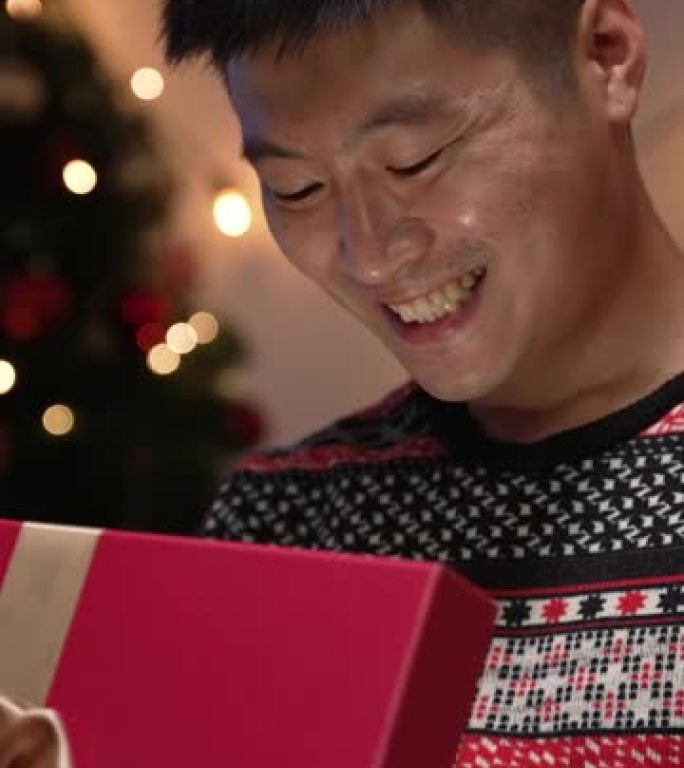 垂直屏幕: 激动的亚洲男子微笑着，握紧拳头欢呼，他在节日的家庭内部接受梦想的礼物，晚上有圣诞节装饰