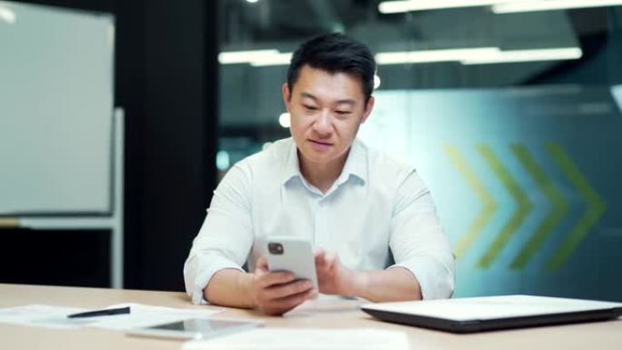 快乐的亚洲上班族坐在办公室的电脑桌前使用手机。开朗的商人企业家员工。