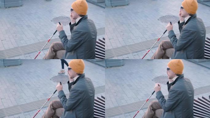 年轻的盲人，智能手机坐在城市公园的长凳上，打电话给