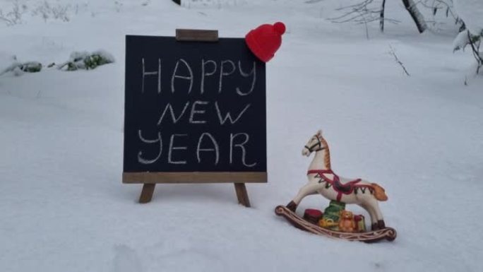 新年快乐祝黑板上的题字文字和圣诞装饰玩具马户外雪地上的礼物。节日庆典