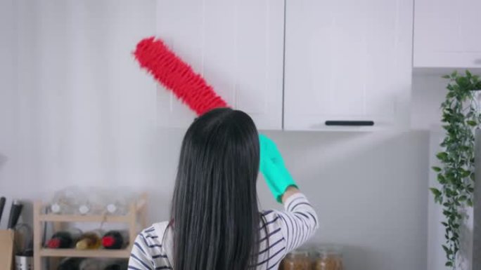 年轻的亚洲妇女用掸子打扫家。漂亮的清洁服务工人家政和整理厨房。家务和家务概念