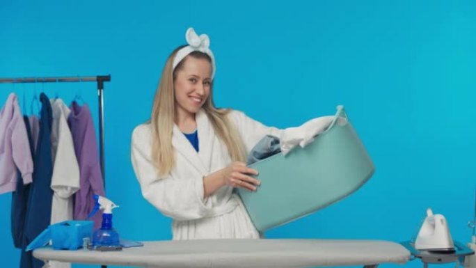 微笑的女人站在工作室碗里的蓝色背景上，衣服放在熨衣板上，打扫卫生，由迷人的金发女人做家务