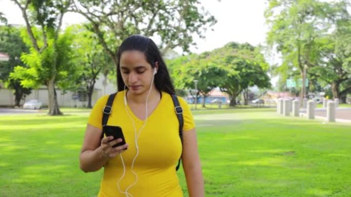 一名西班牙裔妇女带着白色有线耳塞和一部智能手机在巴拿马公园散步的高清视频