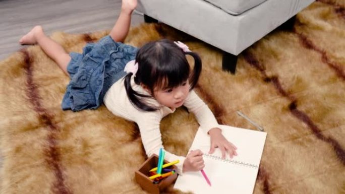 4K，一个可爱的亚洲女孩躺在房子地板上的地毯上，手里拿着蜡笔，高兴地在房子客厅的笔记本上画画。家庭幸
