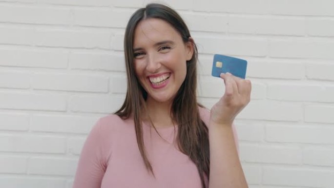 年轻美丽的西班牙裔妇女微笑着自信地拿着信用卡在孤立的白砖背景上