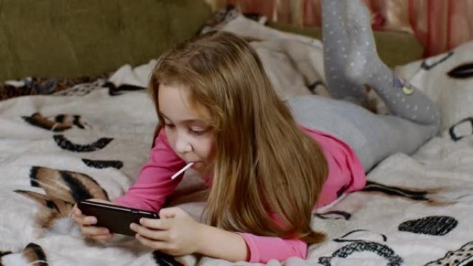 可爱的金发女孩，嘴里叼着棒棒糖，摇晃着双腿，通过点击智能手机屏幕热情地玩耍，躺在铺有轻毯子的沙发上。
