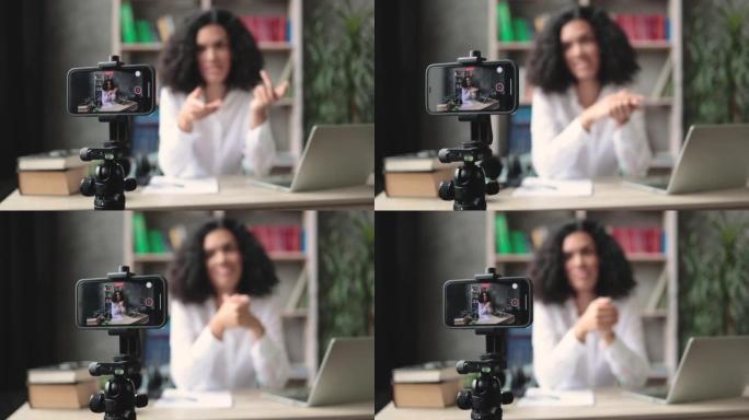 模糊背景的漂亮的跨国女人，黑发坐在办公桌前拍摄视频博客。