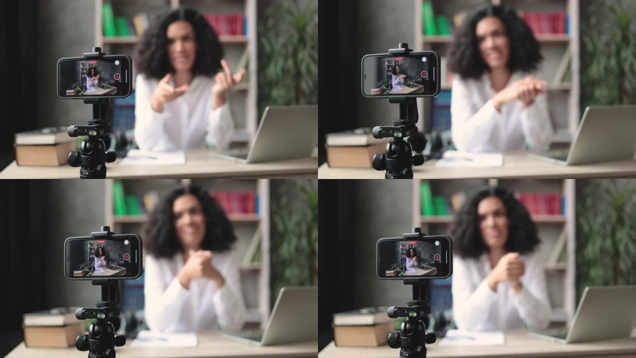 模糊背景的漂亮的跨国女人，黑发坐在办公桌前拍摄视频博客。