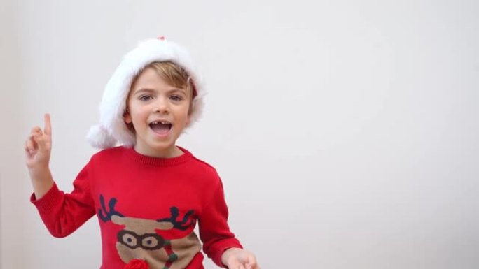 兴奋有趣的笑的小女孩戴着红色的圣诞老人帽子和圣诞驯鹿毛衣在白色背景下快乐地在镜头前跳舞。新年礼物购物