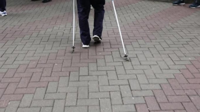 一位老人的腿在城市公园的人行道上一步步行走的后视图。老年体育活动的概念。使用登山杖进行步行支撑。