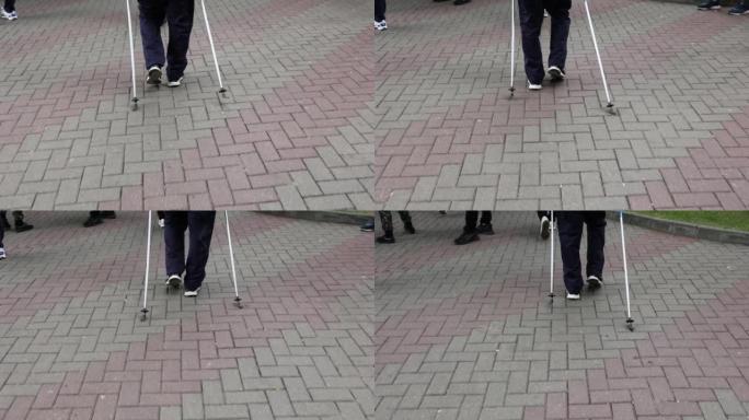 一位老人的腿在城市公园的人行道上一步步行走的后视图。老年体育活动的概念。使用登山杖进行步行支撑。
