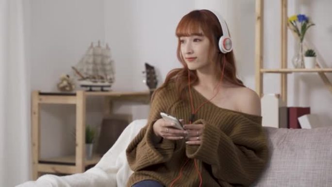 背景明亮舒适的客厅。手持智能手机的快乐中国女士一边听着耳机的轻音乐一边摇晃着身体。