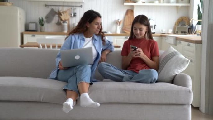 好奇的妈妈女人拿着笔记本电脑看着女儿的手机坐在沙发上，旁边是微笑的少女