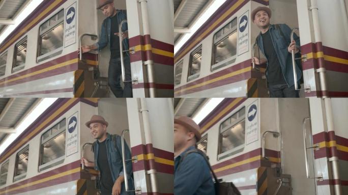快乐的亚洲英俊的背包客男子戴着帽子，牛仔夹克和背包微笑到达目的地时，他抓住火车栏杆以确保安全，并走出