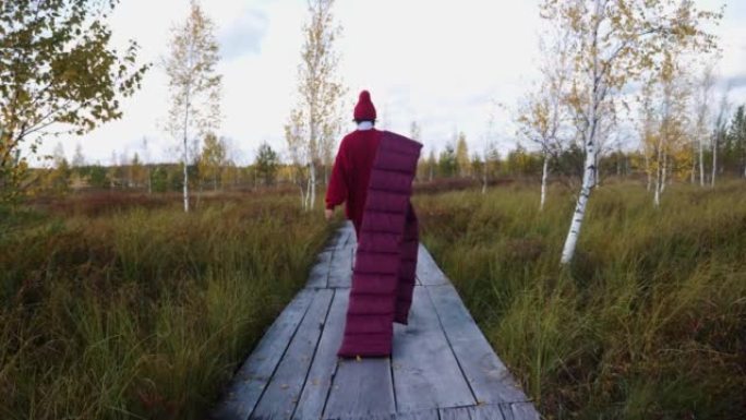 在白俄罗斯的叶尔尼亚，一名穿着红毛衣、戴着帽子、戴着围巾的时尚女子在秋天的沼泽中沿着木板铺成的小径走