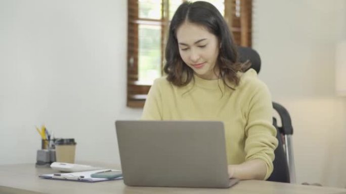 在笔记本电脑上工作的亚洲女性看着笔记本电脑屏幕，通过互联网通信系统在家放松工作