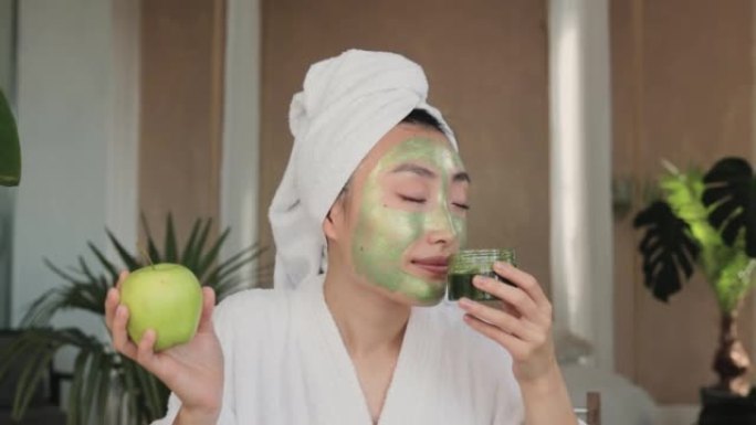 愉快的亚洲女人，头上有浴巾，绿色面膜，拿着苹果和玻璃器皿