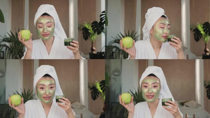 愉快的亚洲女人，头上有浴巾，绿色面膜，拿着苹果和玻璃器皿