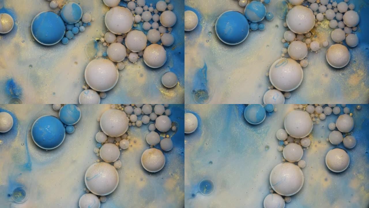 彩色白色金色和蓝色油漆气泡散布在蓝色水彩表面上，并在抽象设计中混合。闪闪发光的颗粒，墨球滴和混合。运