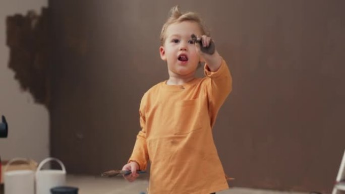 微笑的两岁男孩站在房间里，手里拿着刷子。金发男孩穿着橙色毛衣，手臂和毛衣上涂有棕色油漆。后面是涂漆的
