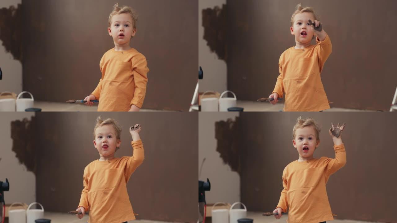 微笑的两岁男孩站在房间里，手里拿着刷子。金发男孩穿着橙色毛衣，手臂和毛衣上涂有棕色油漆。后面是涂漆的