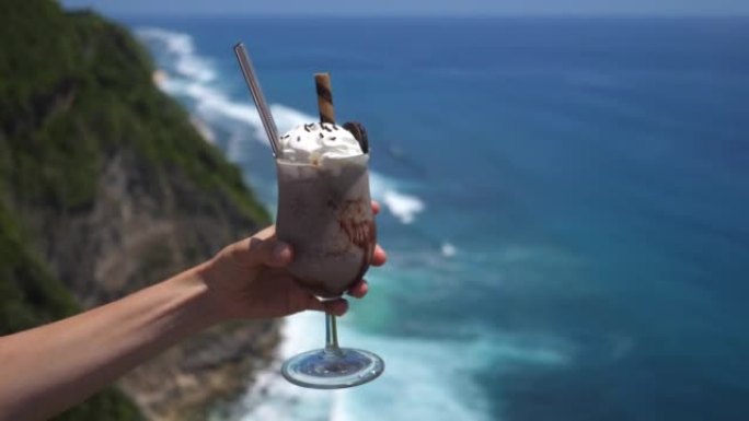 纯素食巧克力奶昔，上面放着鲜奶油，手里拿着奥利奥饼干，在美丽的蓝色海洋和翠绿的悬崖背景下。