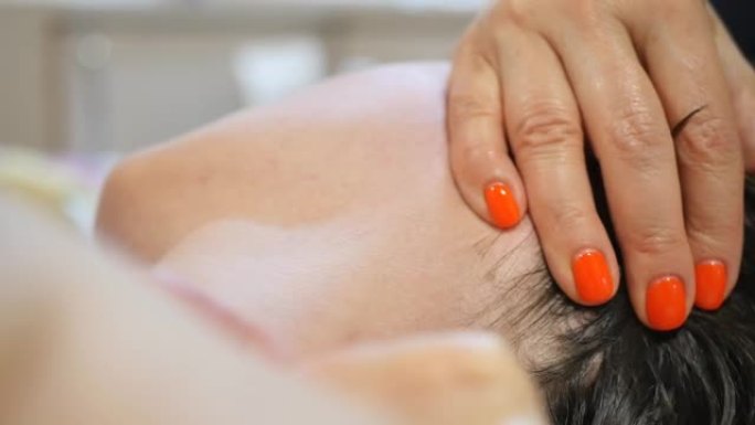 特写按摩女性颈部和枕部区域在按摩面部和头部的过程中，用美容师按摩师的手与红色修指甲。面部和头皮护理