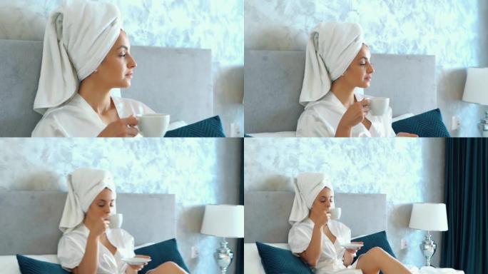 白种人漂亮的女人穿着长袍，头上戴着白毛巾，躺在床上，洗完澡后在卧室里喝咖啡。年轻的女性早上在家里的床