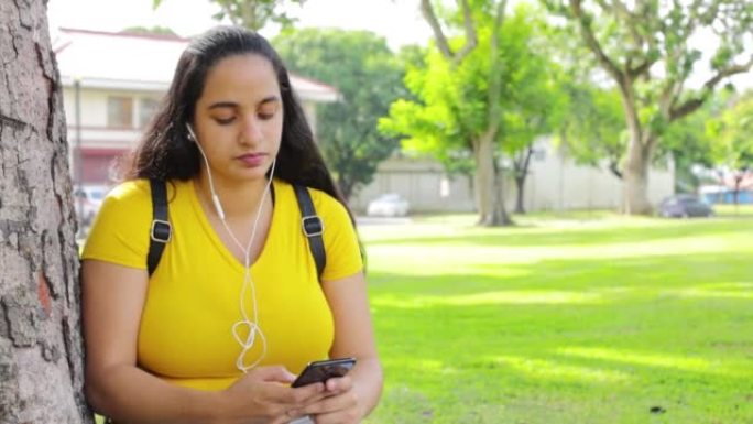 一个带耳塞和智能手机的西班牙裔女孩在公园听音乐和发短信的高清视频