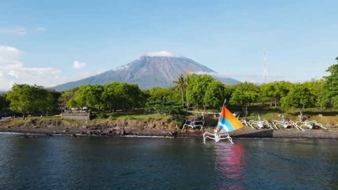 传统巴厘岛渔船的彩帆驶向沿海海滩，与高耸的阿贡火山相形见war。