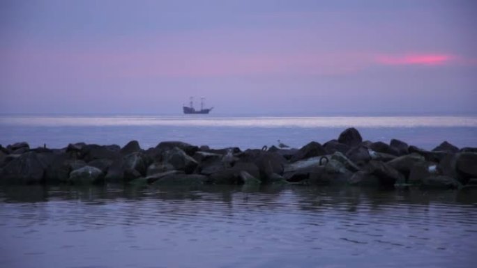 波罗的海。蓝色小时。前景石质防波堤上的海鸥。背景中风格化为海盗的船。紫色。波兰达尔沃沃