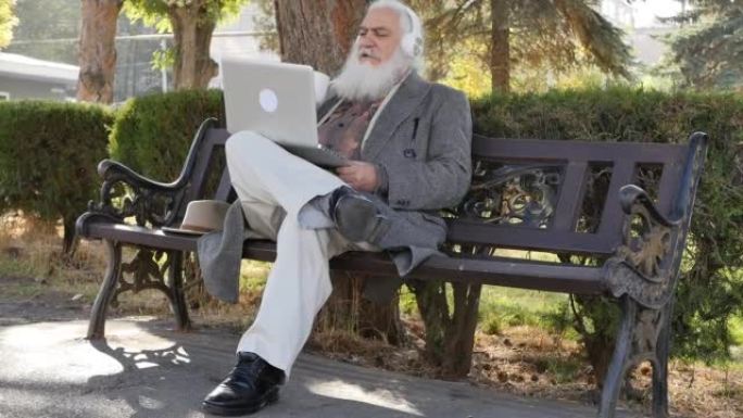 在秋天的公园里，留着蓬松的灰色胡须的有魅力的高级男子坐在长凳上，用笔记本电脑和白色耳机通过视频链接交