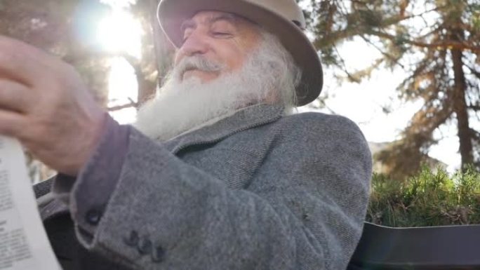 一个穿着灰色外套，戴着帽子的大胡子的老人在一个阳光明媚的早晨坐在长凳上的公园里看报纸，为报纸的好消息