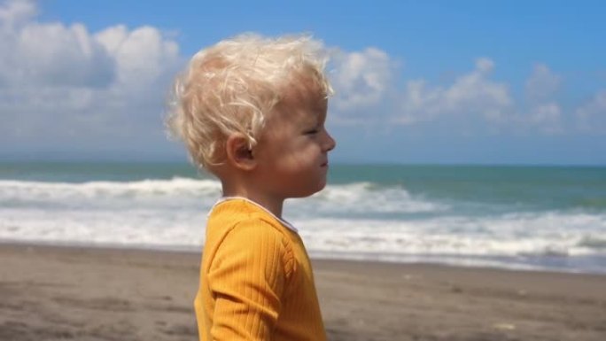 穿着亮黄色上衣的金发蹒跚学步的肖像，在沙滩上表达了不满。与孩子们在海上度假。在慢动作中，海浪在岸上坠
