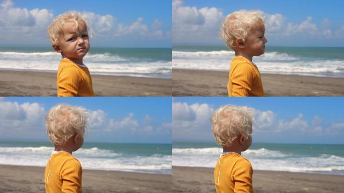 穿着亮黄色上衣的金发蹒跚学步的肖像，在沙滩上表达了不满。与孩子们在海上度假。在慢动作中，海浪在岸上坠