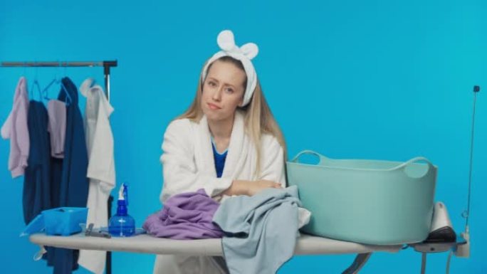 早晨，女人为洗衣碗中要熨烫的衣服感到难过。一个女孩站在工作室的蓝色背景上，用乏力擦拭额头