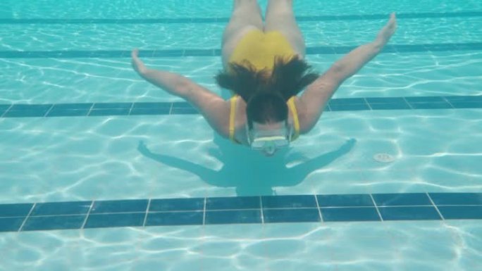 一个穿着黄色泳衣和面具的胖乎乎的女孩从镜头前的水下弹出。游泳池和水下相机。慢动作