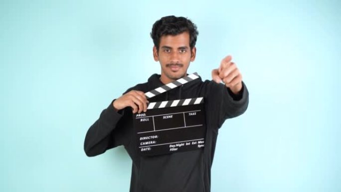 年轻的亚洲印度男子站着拿着隔板，用食指指着相机，拍片用拍板，孤立在彩色背景工作室肖像上