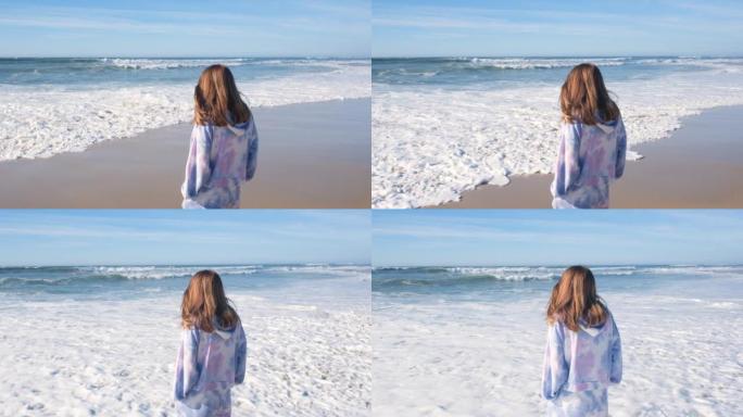 穿着tieday track suite的年轻美女看着大西洋海岸和泡沫的海浪。海滩，蓝天和向远处望去
