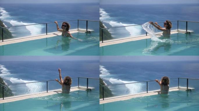 高加索女孩在俯瞰大海的无边泳池里泼水。在蓝色海洋岸边的一家昂贵的酒店里享受奢华的假期。水融合效应。