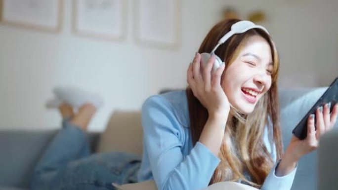 快乐的年轻亚洲女人在家放松。女性微笑坐在沙发上，拿着移动智能手机。女孩用视频打电话给朋友。听音乐
